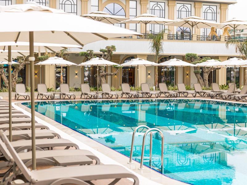 Área de piscina con tumbonas y sombrillas frente a un edificio de hotel en un día soleado.