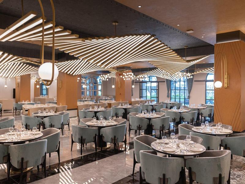 Restaurante elegante del hotel con mesas redondas, sillas claras y diseño moderno de luces en el techo.