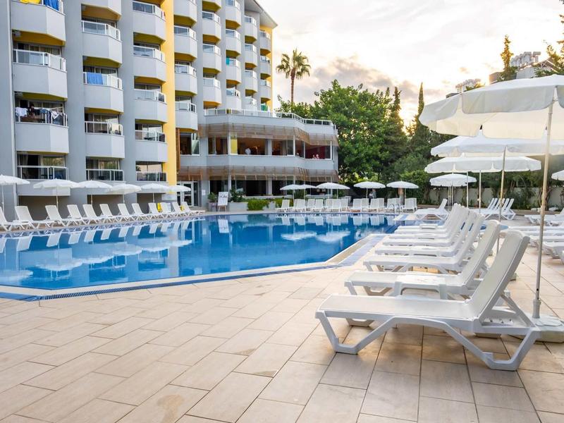 Empty hotel pool area with white lounge chairs under umbrellas and multiple balconies.