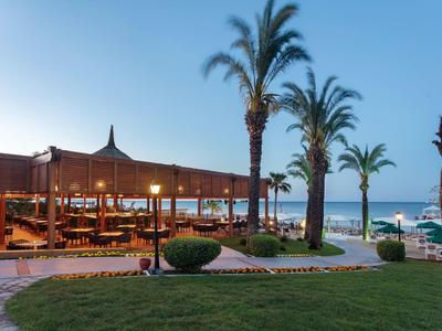 Strandrestaurant mit Holzterrasse, Palmen, grünem Rasen und blauem Himmel am Meer im Hintergrund.