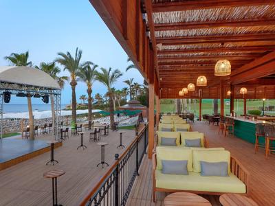 Strandbar mit Holzterrasse, gelben Sofas, Palmen und einer Bühne am Meer bei klarem Himmel.