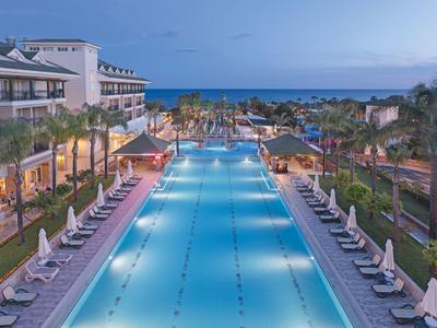 Großer beleuchteter Pool mit Liegen und Palmen, Hotelgebäude links, Meer im Hintergrund.