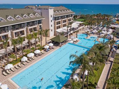 Großer blauer Pool mit Liegestühlen, Palmen und einem Hotelgebäude neben dem Meer.