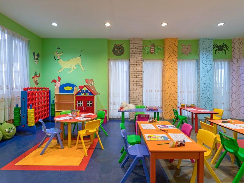 Bunter Kindergartenraum mit bunten Tischen, Stühlen, Spielzeugen und großen Fenstern mit Vorhängen.