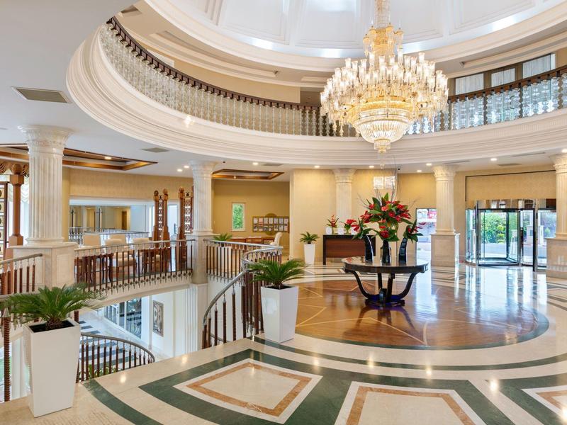 Großer Hotellobby mit Marmorboden, Kronleuchter, runder Treppe und Blumenarrangements.