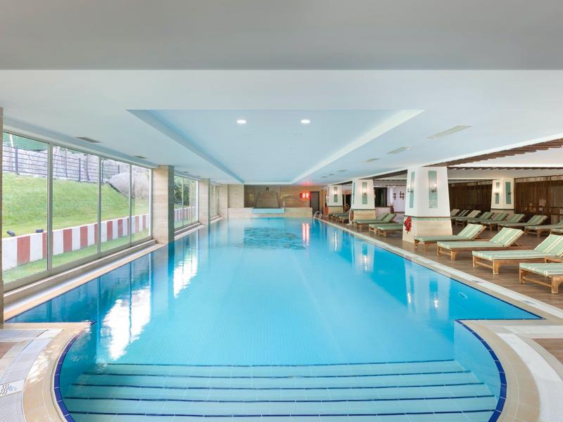 Helles Hallenbad mit blauen Fliesen, Liegen und großem Fenster zur Außenanlage.