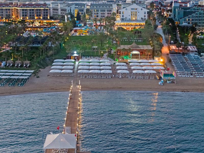 Strand mit Steg, Sonnenschirmen und Hotelanlage im Hintergrund bei Sonnenuntergang.