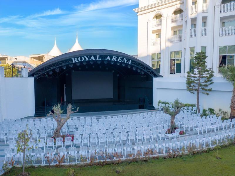 Venkovní jeviště s bílými židlemi před bílou budovou pod modrou oblohou.