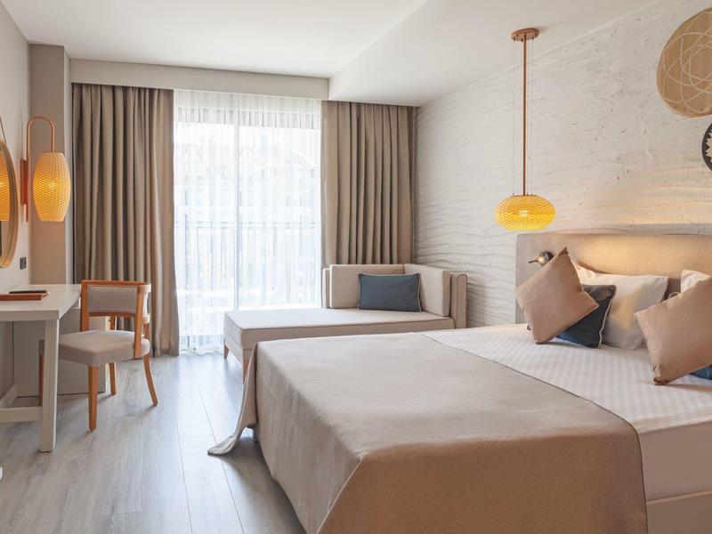 Moderní hotelový pokoj s velkou postelí, pracovním stolem, pohovkou a světlými závěsy.