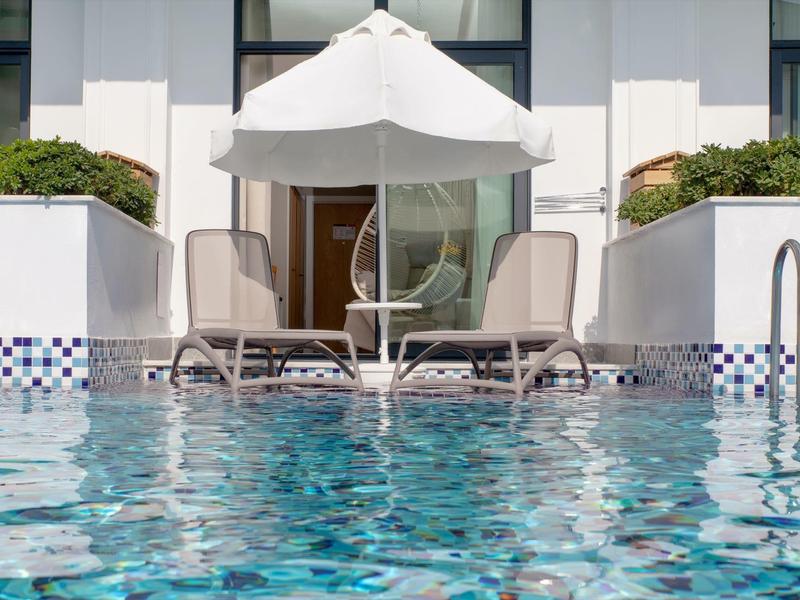 Dva lehátka s bílým slunečníkem vedle bazénu před moderní budovou.