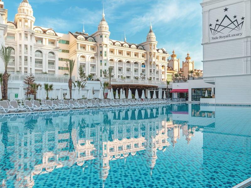 Velký venkovní bazén před elegantní vícepodlažní hotelovou budovou za slunečného dne.