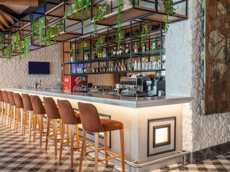 Moderní hotelový bar s vysokými koženými stoličkami a dekorativními rostlinami nad pultem.