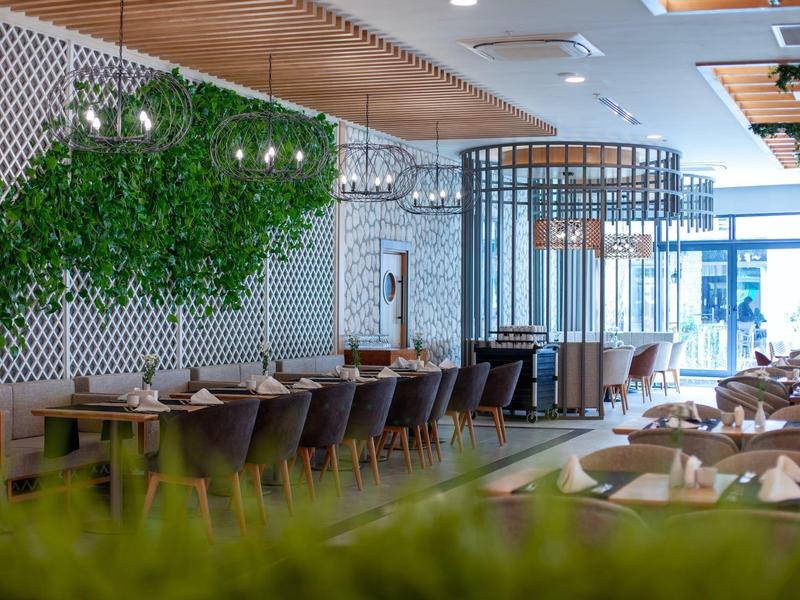 Moderní hotelová restaurace s dřevěnými stoly, zelenými rostlinami a dostatkem přirozeného světla.