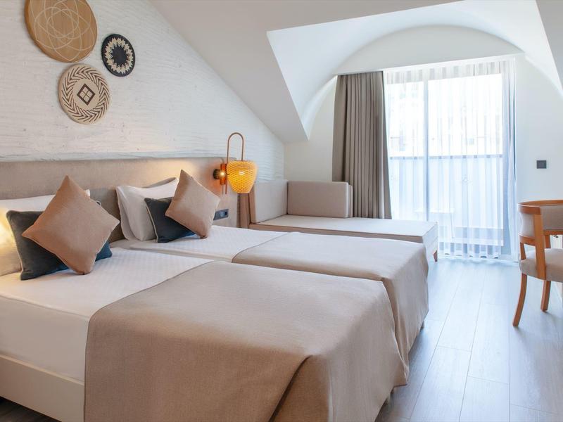 Světý hotelový pokoj s dvěma jednolůžky, béžovými přikrývkami a židlemi u okna.