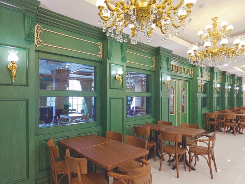 Elegantní jídelní prostor se zelenými nástěnnými panely, dřevěnými stoly a židlemi, osvětlený lustry.