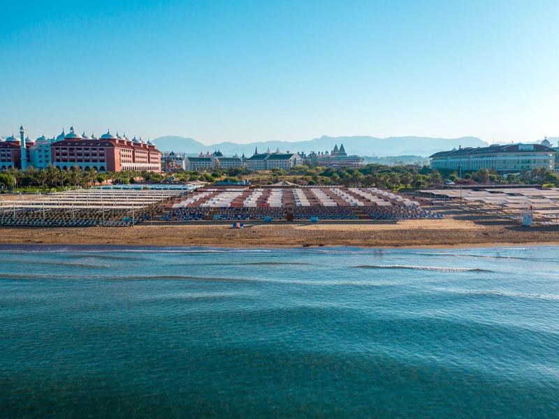 Přímořský výhled s písečnou pláží, řadami lehátek a velkou budovou v pozadí.
