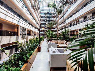 Lobby di hotel moderna con alti soffitti in vetro, piante e aree salotto accoglienti.