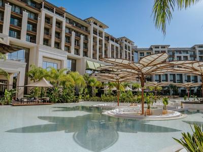 Hotel con arquitectura moderna, gran piscina y sombrillas bajo cielo azul.