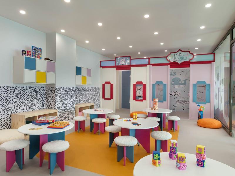 Área infantil moderna y luminosa del hotel con mesas coloridas, cojines y juegos de pared.