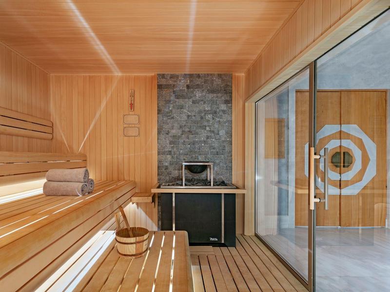Sauna luminosa con revestimiento de madera, bancos, cubo y toallas junto a una puerta de vidrio.