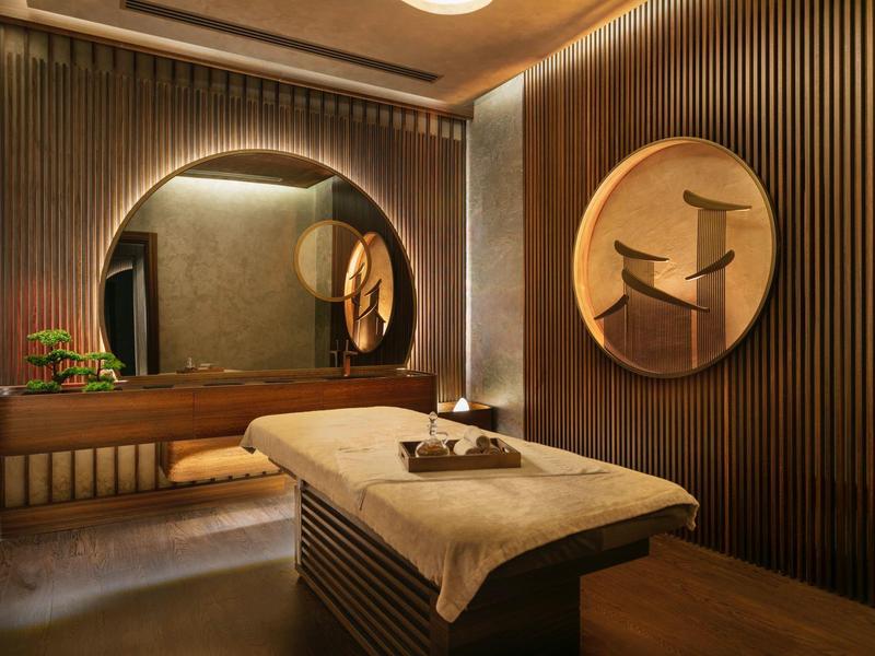 Sala de bienestar elegante con camilla de masaje, espejo redondo grande y decoración en madera oscura.