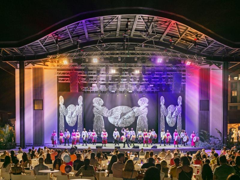 Escenario al aire libre con iluminación colorida y grupo de baile frente a público de noche.