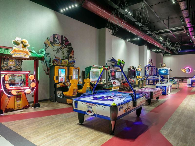 Sala de juegos moderna con máquinas arcade y mesas de air hockey bajo luces coloridas.