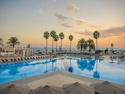 Piscine extérieure avec chaises longues et palmiers au coucher du soleil, vue sur la mer.