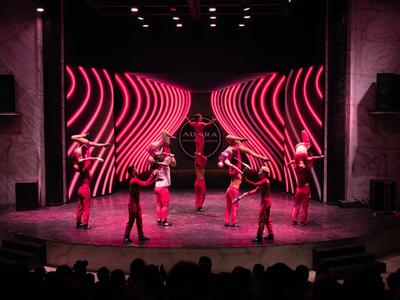 Palco con cinque ballerini in abiti rossi davanti a uno sfondo con linee ondulate rosa luminose