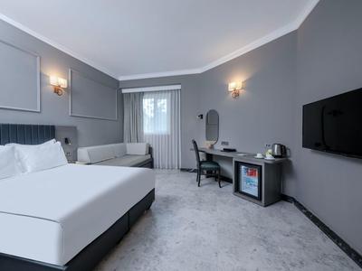 Chambre d'hôtel grise avec lit double, canapé, bureau et TV murale.