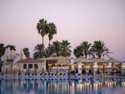 Piscine calme devant un hôtel avec des chaises longues et des palmiers au coucher du soleil.