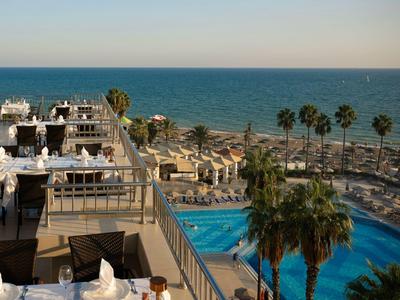Terrasse d'hôtel avec piscine, palmiers et vue sur la mer calme sous un ciel clair.