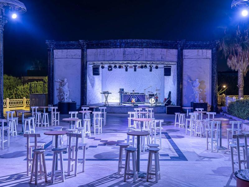 Terrasse extérieure avec tables hautes et scène éclairée pour événements nocturnes.