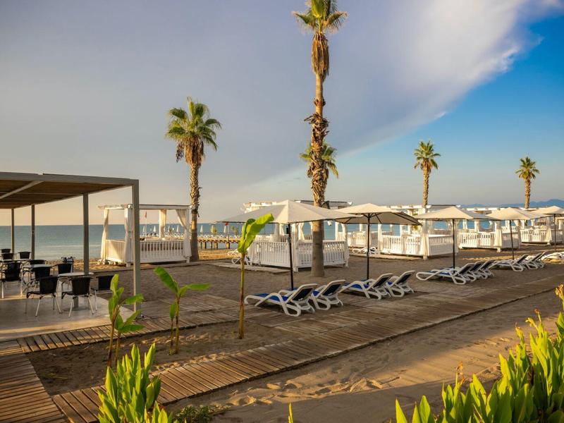 Plage avec chaises longues, parasols blancs et palmiers sous un ciel bleu avec nuages.