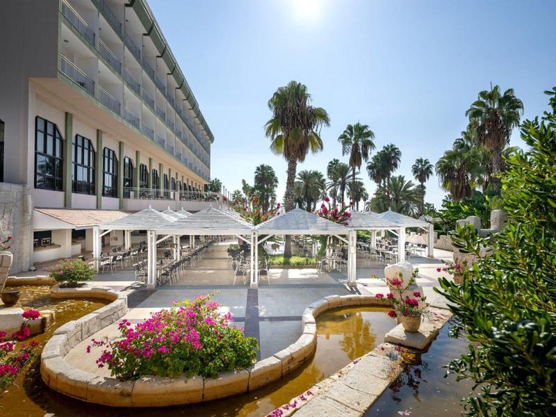 Hôtel avec une fontaine entourée de fleurs et de palmiers sous un ciel ensoleillé.