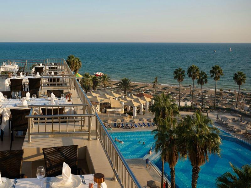 Terrasse d'hôtel avec piscine, palmiers et vue sur la mer calme sous un ciel clair.