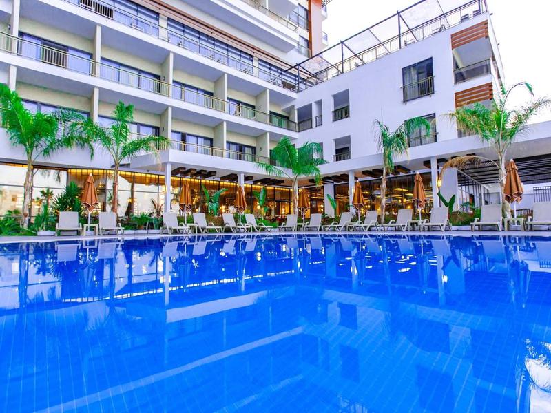 Espace piscine d'hôtel moderne avec parasols, palmiers et bâtiment à plusieurs étages en arrière-plan.