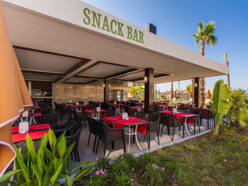 Snack-bar couvert avec des tables et chaises rouges dans un cadre de jardin sous un ciel bleu.