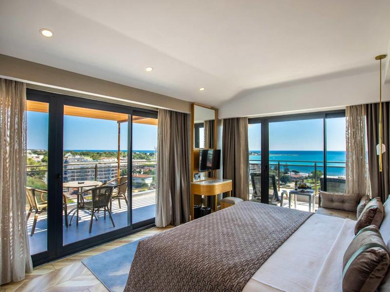 Chambre d'hôtel moderne avec grand lit, balcon et vue sur la mer au coucher du soleil.