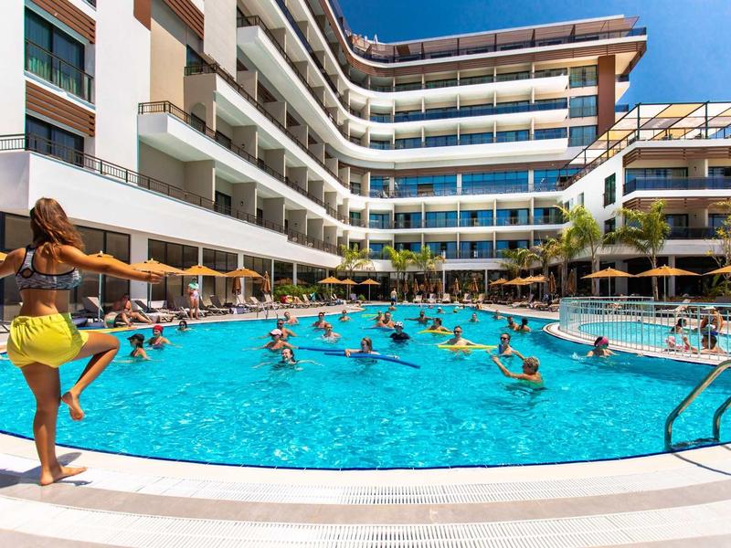 Des personnes participent à un cours d'aquafitness dans une piscine extérieure d'un hôtel moderne par une journée ensoleillée.
