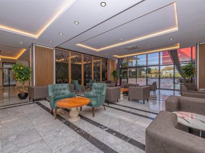 Moderní hotelový salon se zelenými křesly, dřevěnými stolky a velkými okny.