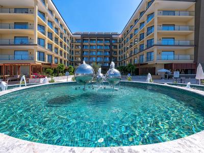 Großer runder Pool mit springenden Wasserspielen vor modernen mehrstöckigen Gebäuden bei klarem Wetter.