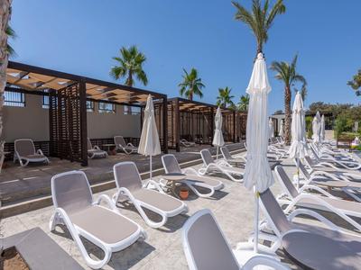 Weiße Liegestühle und geschlossene Sonnenschirme auf einer Terrasse unter blauem Himmel neben einer Steinfassade.