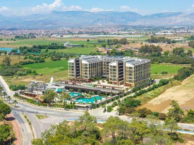 Moderní hotel s bazénem obklopený zelenými poli a horami v pozadí.