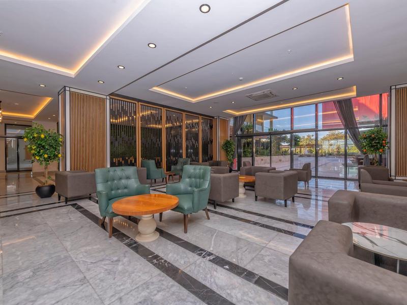 Moderní hotelový salon se zelenými křesly, dřevěnými stolky a velkými okny.