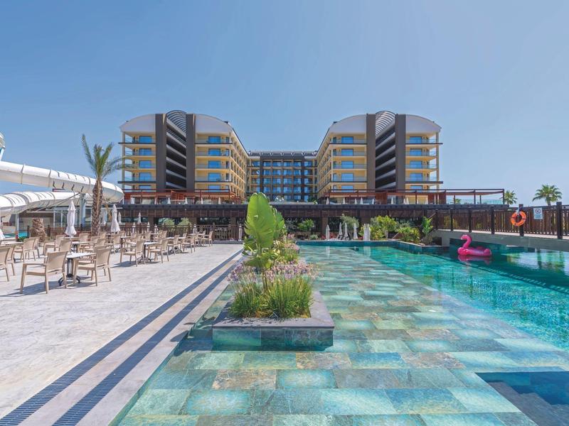 Tyrkysový venkovní bazén s moderním hotelem v pozadí pod světle modrou oblohou.