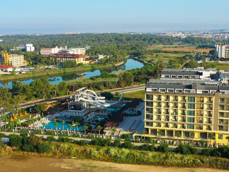 Moderní hotel u řeky s bazénem, obklopený zelení a obytnými čtvrtěmi.