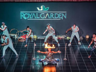 Eine dynamische Tanzgruppe führt vor einer "Royal Garden"-Leinwand auf einem glänzenden schwarzen Boden mit Feuer auf.