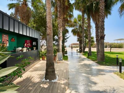 Un paseo soleado bordeado de palmeras junto a un hotel y un bar al aire libre.