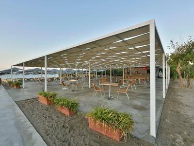Offene Pergola mit Tischen und Stühlen entlang eines Küstenwegs bei klarem Himmel.